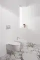 Weißes Wand-Bidet und WC von Ideal Standard in einem modernen Badezimmer mit Marmorfußboden, hellen Wänden und einem Fenster mit Handtüchern.